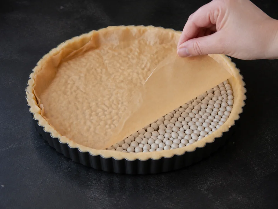 Quicheteig in einer 26-Zentimeter-Tarteform wird mit Backpapier und Blindbackkugeln vorgebacken