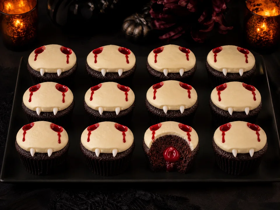 Zwölf Vampir-Cupcakes mit heller Creme, roten Bissspuren und Zuckerzähnen auf einer schwarzen Platte